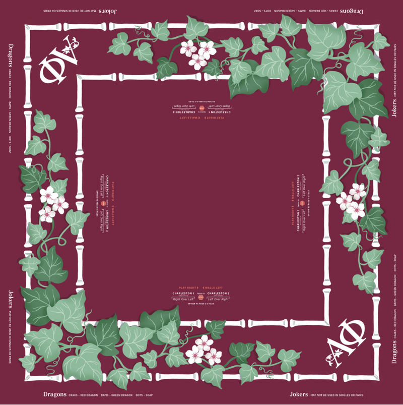 Alpha Phi MIGTM Mahjong Mat design with a deep burgundy background, featuring a white bamboo-style border decorated with green ivy leaves and clusters of light pink flowers. The corners display the stylized 'DA' logo for Dragons Askari, with text at the top and bottom detailing the brand and collection.